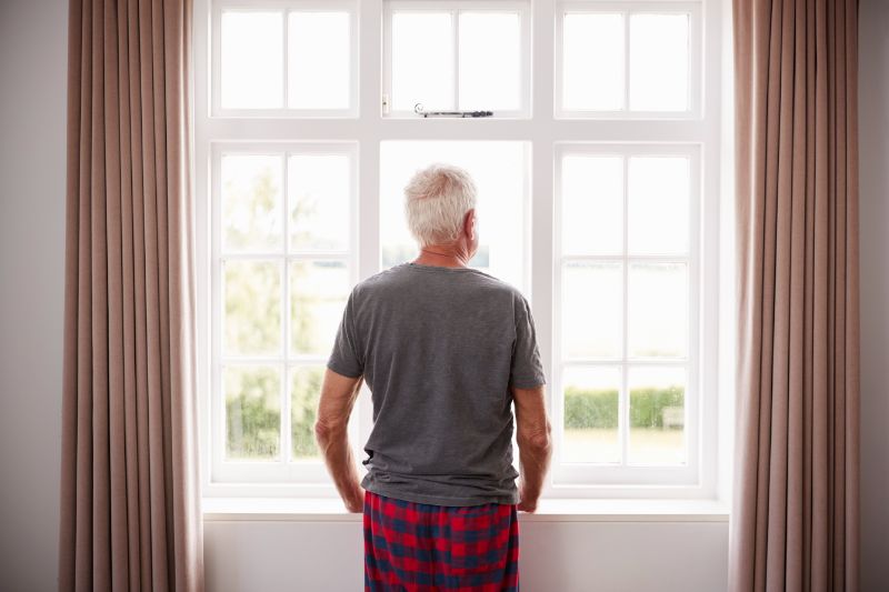 Rear View Of Senior Man In Pajamas Looking Out Of Bedroom Window